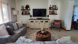 A living room with a grey sofa, a brown ottoman, a television, and a bookshelf with decorative items. - Charming Nauvoo Farmhouse - 1 mile from Nauvoo Temple (Nauvoo)