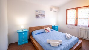 A bedroom with a wooden bed, blue bedding, a blue nightstand with a lamp, a small bedside table, and a window with curtains. - S'Omu and Assunta 1P by All4sardinia  (Cardedu)