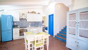 A compact kitchen with white cabinets, a blue refrigerator, and a dining area with a table and chairs. - S'Omu and Assunta 1P by All4sardinia  (Cardedu)