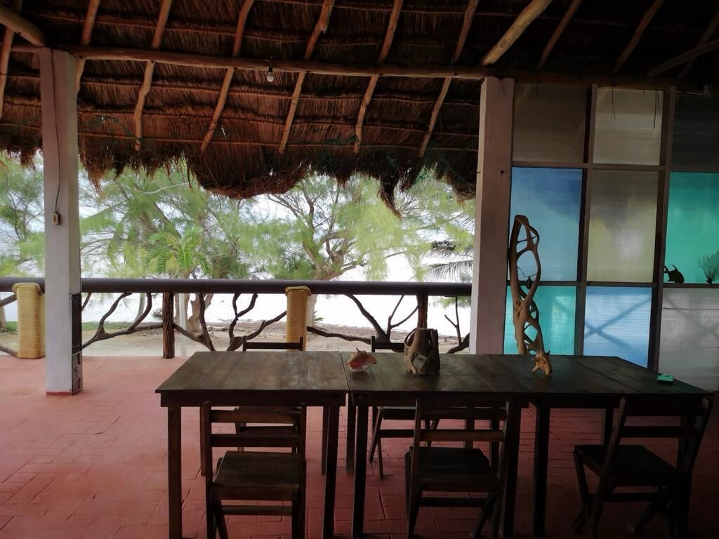 A covered outdoor seating area with a wooden table and chairs, a thatched roof, and a view of palm trees.