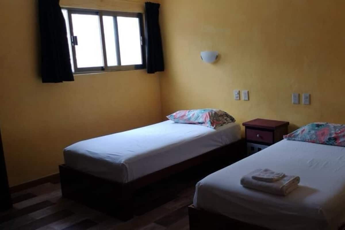 Una habitación con dos camas, cada una con una almohada y una mesita de noche. Hay una ventana con cortinas, y las paredes son de color amarillo.