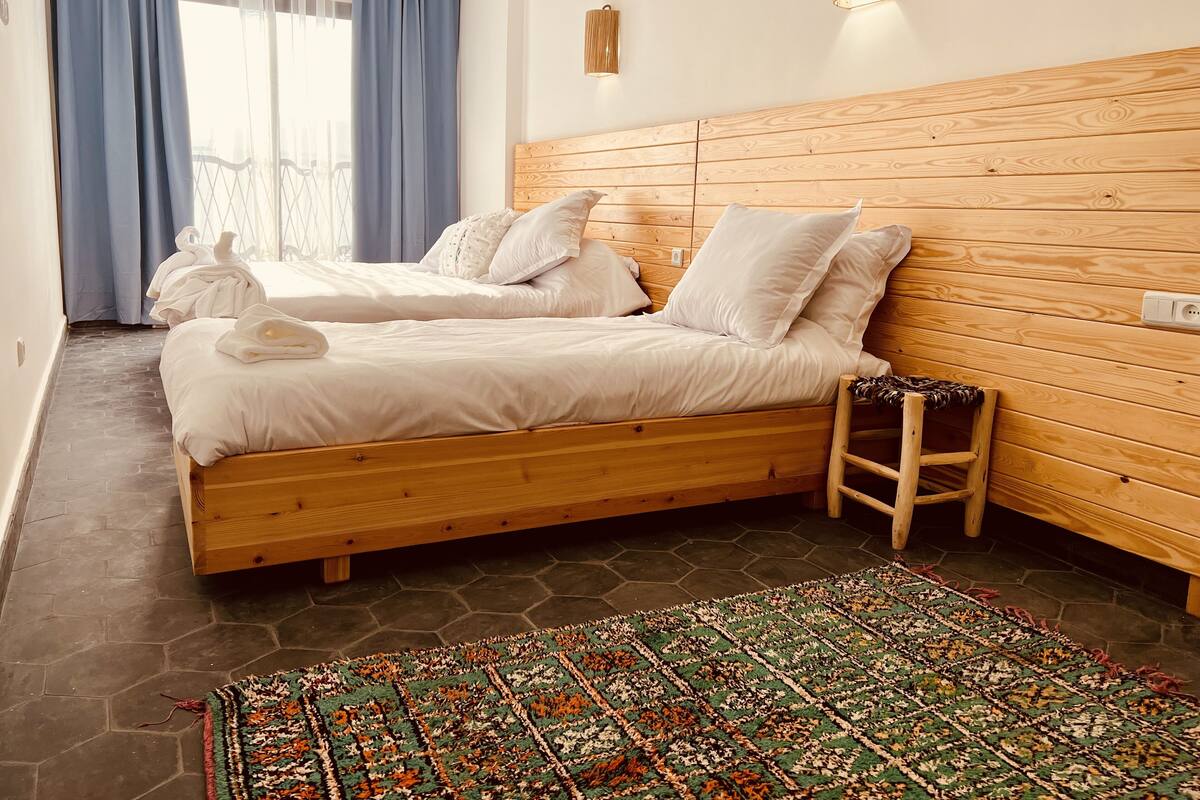 Un lit double avec du linge de lit blanc, une tête de lit en bois et un petit tabouret en bois.