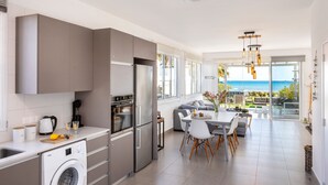 A modern kitchen with a washing machine, a dining area with a table and chairs, and a view of the sea through large windows. - Ayia Thekla Seafront Villa Dagi (Ayia Napa)
