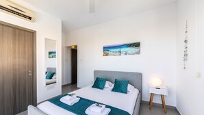 A modern bedroom with a bed, a nightstand, a wall-mounted air conditioner, a door, and a painting of a coastal scene. - Ayia Thekla Seafront Villa Dagi (Ayia Napa)