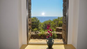 Ein Zimmer mit Blick auf das Meer durch ein steingerahmtes Fenster, eine Vase mit Blumen auf einem Tisch.