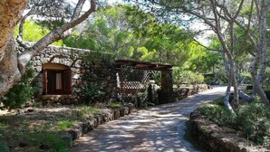 Una cabaña de piedra con puerta de madera y un pequeño porche, rodeada de árboles y un camino pavimentado.