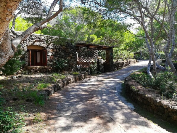 A stone cottage with a wooden door and a small porch, surrounded by trees and a paved path.