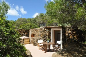 A rustic stone building with a wooden door, a small table, and two chairs in a natural setting.