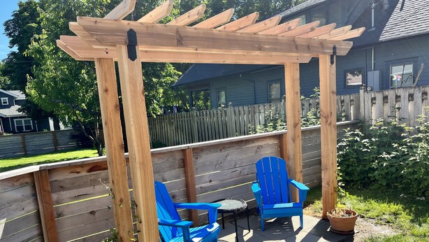 En pergola i træ med to blå stole og et lille bord, placeret på en terrasse med et træhegn og grøn bevoksning i baggrunden.