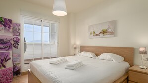 Una cama bien hecha con sábanas y toallas blancas, un cabecero de madera, una mesita de noche con lámpara, y un ventanal con vistas al exterior.
