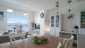 Un comedor con mesa de madera, sillas blancas, un jarrón con plantas y vistas amplias a través de ventanas grandes.