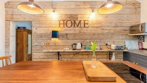 A modern kitchen with wooden walls, a wooden dining table, pendant lights, and a visible 'HOME' sign. - The Cow Shed—Luxury Barn Moments From Narberth (Narberth)