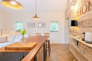 Een moderne keuken met een houten aanrecht, barstoelen en een inbouwoven. Er hangen ingelijste foto's aan de muur en er is een raam met zonwering.