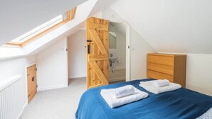 Ein Schlafzimmer mit schrägen Wänden, einem Oberlicht, einem Holzschrank, einem Bett mit blauer Bettwäsche und einem Badezimmer mit Waschbecken und Spiegel.