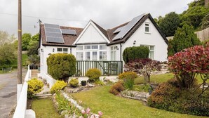 Una casa de dos pisos con paneles solares en el techo, un jardín bien cuidado con diversas plantas y flores, y una cerca blanca de piquete.