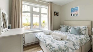 Un dormitorio con un ventanal grande, una cama con ropa de cama floral, una mesita de noche y un cuadro en la pared.