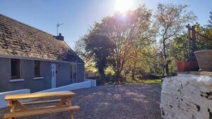 Eine gepflasterte Terrasse mit einem Holztisch, einer weißen Mauer und einem Haus mit grauer Fassade und geneigtem Dach.