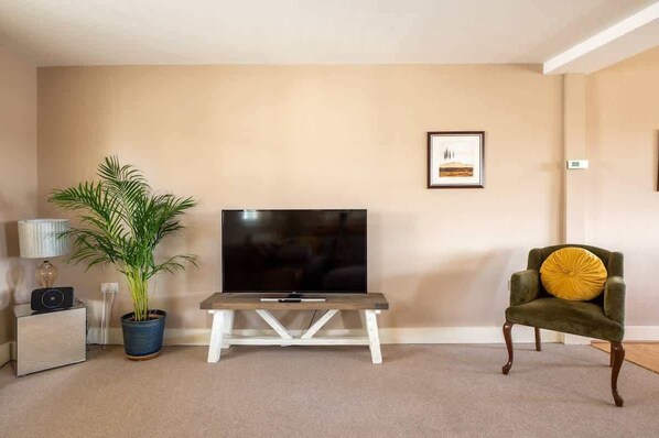 A living room with a flat-screen TV on a wooden stand, a green armchair with a yellow cushion, a potted plant, and a framed picture on the wall.