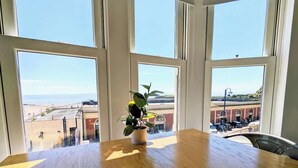 Una mesa de madera con una planta en maceta en el centro, amplias ventanas con vistas a una playa y edificios.