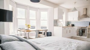 Habitación de hotel moderna con una cama grande, un comedor con mesa y sillas, una zona de cocina y vistas amplias a través de grandes ventanales.