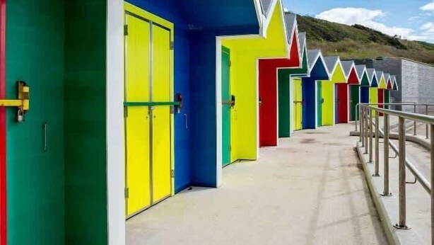 Una hilera de cabañas de playa de colores brillantes, con puertas y ventanas, un paseo de hormigón y una barandilla de metal.
