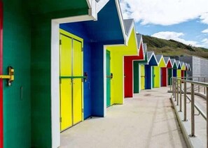 A row of brightly colored beach huts with doors and windows, a concrete walkway, and a metal railing. - Barry Island Beachfront Getaway + Private Parking (Vale of Glamorgan)