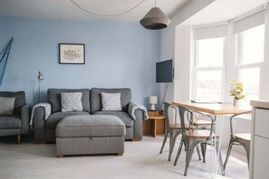 Uma sala de estar moderna com um sofá cinzento, uma mesa de jantar com cadeiras, uma televisão e um quadro na parede.