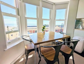 Quarto de canto com uma mesa de madeira e quatro cadeiras, janelas amplas com vista para a praia e uma planta em vaso sobre a mesa.
