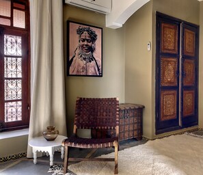 A room with a woven chair, a small table with a bowl, a wall-mounted picture, and a large ornate door. - Entire Riad Dar Chacha - privatised only for you (Marrakesh)