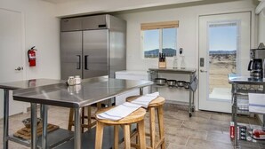 A modern kitchen with stainless steel appliances, a large island, bar stools, and a view of the desert through the door.