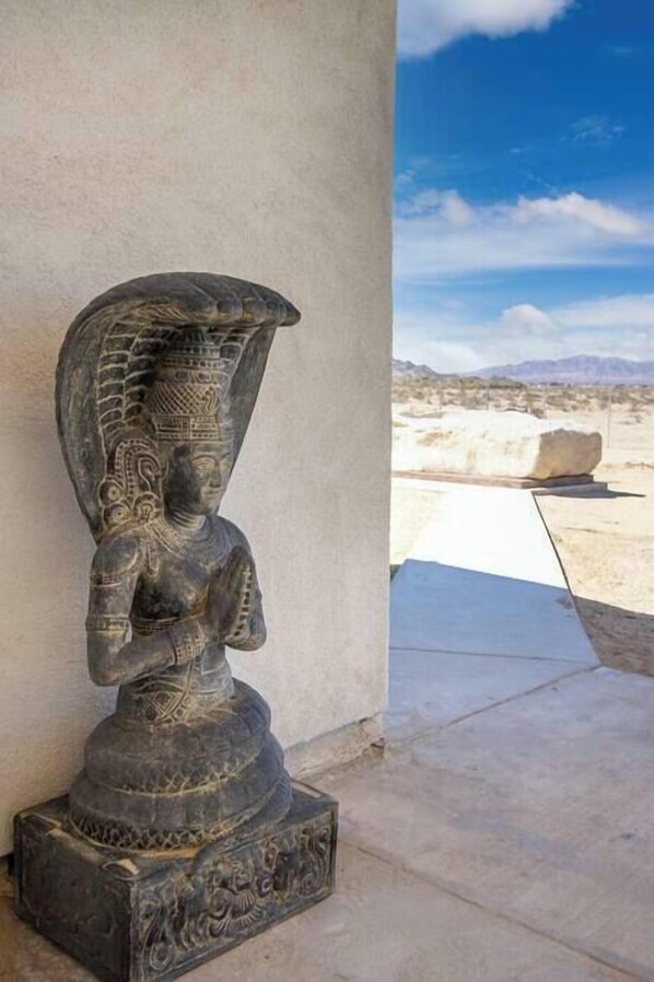 A stone sculpture of a seated figure with intricate patterns, placed against a plain wall with a clear sky and distant mountains in the background.