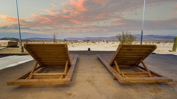 Two wooden lounge chairs facing a desert landscape with mountains in the distance and a colorful sky.