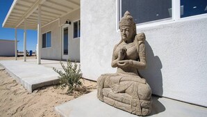 A stone statue of a seated figure next to a building with a covered porch and a window.