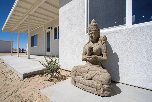 A stone statue of a seated figure next to a building with a covered porch and a window.