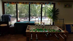 Una habitación con mesa de fútbol, mesa de billar y vistas a un patio trasero con piscina y mobiliario de jardín.