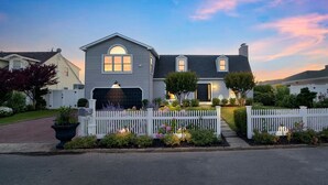 Una casa de dos pisos con garaje, rodeada por una cerca blanca de piquetes y un jardín bien cuidado.