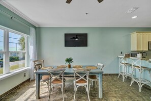 A dining area with a table set for four, surrounded by white wicker chairs. A flat-screen TV is mounted on the wall above the table.
