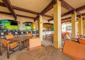 Un área de comedor al aire libre con mesas y sillas de madera, una barra con mostrador verde y una cocina al fondo.