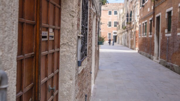A narrow street with buildings on either side, featuring a wooden door and a window with a metal grate. - CA SORANZO (Venice)