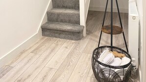 Un escalier avec des marches recouvertes de moquette grise, des murs blancs et une main courante en bois. Un lampadaire noir avec un pied en bois est placé près de l’escalier.
