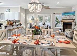 A dining area with a large table set for a meal, a chandelier, and a television in the background. - Tangerine Cove-5 Min 2 Beach-Golf Cart Deals (Destin)