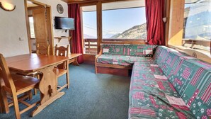 Een gezellig interieur van een chalet met een houten eettafel, een bank met een kleurrijke quilt en een televisie aan de muur.
