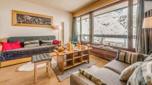 Een gezellige woonkamer met een bank, een bijzettafeltje met ontbijtartikelen en een uitzicht op besneeuwde bergen door grote ramen.