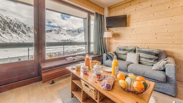 Een gezellige kamer met houten details, een bank, een tafel met ontbijtartikelen en een groot raam met uitzicht op een besneeuwde berg.