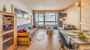 Een gezellige, goed verlichte kamer met houten inrichting, een bank, een eettafel gedekt voor twee personen en een groot raam met uitzicht op besneeuwde bergen.