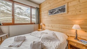 Een hotelkamer met een wand van houten panelen, een bed met witte lakens, twee bedlampen, een raam met uitzicht op besneeuwde bergen en een radiator onder het raam.