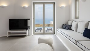 Ein modernes Wohnzimmer mit Fernseher, Sofa, Hocker und Meerblick durch große Glasfenster.