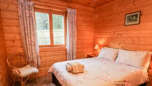 A wooden cabin room with a bed, a chair, a window with curtains, and a framed picture on the wall. - Chloe's Lodge (Pickering)