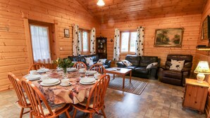 A wooden cabin interior with a dining area, a living room with a sofa and armchairs, and a fireplace. - Chloe's Lodge (Pickering)