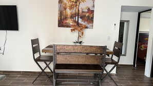 A dining area with a wooden table and chairs, a painting of a tree-lined path, and a potted plant. - Newly renovated centrally located apartment in La Crucecita Huatulco Oaxaca  (La Crucecita)